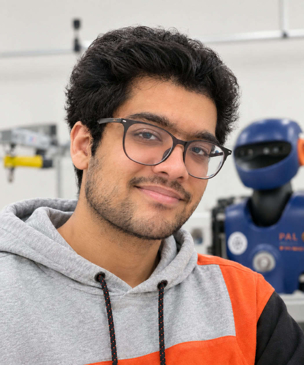 Ayush Deshmukh in a robotics lab, with a blue humanoid robot behind him